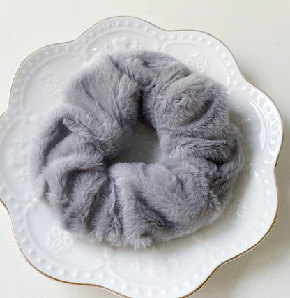 Chouchou en fausse fourrure gris pastel posé sur une assiette blanche décorative, présentation élégante du produit.